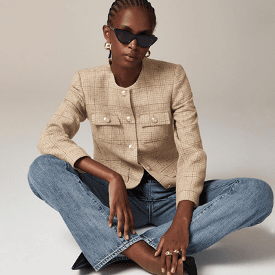 Mulher sentada usa jaqueta bege com bolsos de remendo, abotoado no pescoço; Ela está sentada com maçã cruzada e vestindo jeans e óculos de sol.