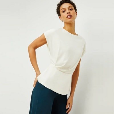 mulher profissional encostada na parede; ela usa um top branco de manga curta da MMLaFleur com detalhes drapeados e calça verde escura.