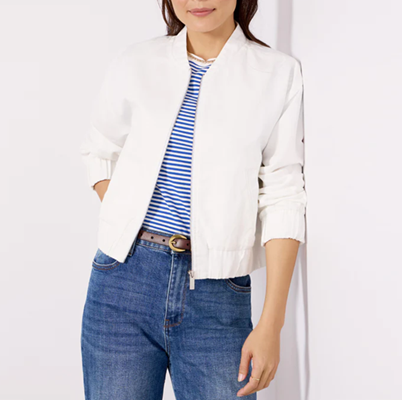 Mulher se inclina contra a parede usando jaqueta de linho branco com mangas empurradas; Ela tem uma camiseta listrada e jeans azul e tem uma mão no bolso