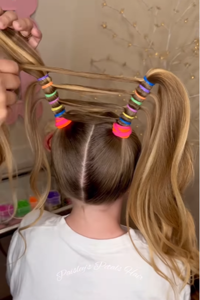 Mulher demonstra tranças espetadas em uma criança usando múltiplos elastics para tornar os tranças rígidos e conectar os dois tranças no meio para uma coroa linear