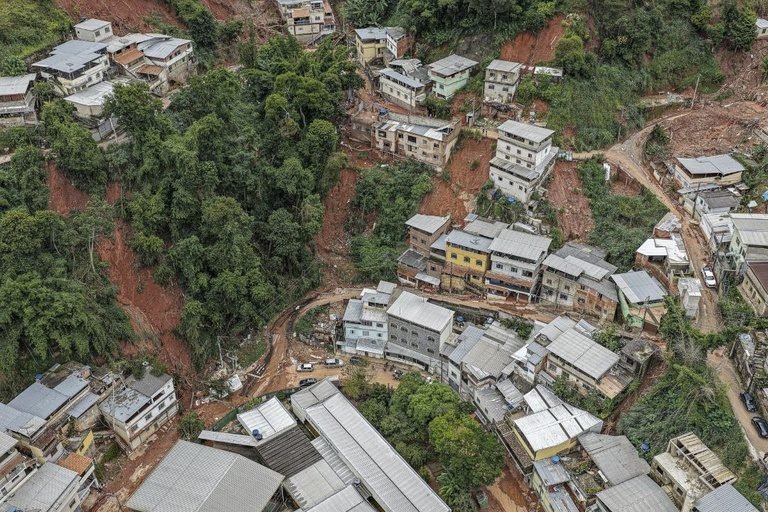 Imagem aérea mostra casas construídas em área de morro, cercadas por vegetação densa.Há trechos de encosta expostos, com solo avermelhado e sinais de deslizamento ou erosão.As moradias ficam próximas a barrancos e vias estreitas em uma região de ocupação irregular.