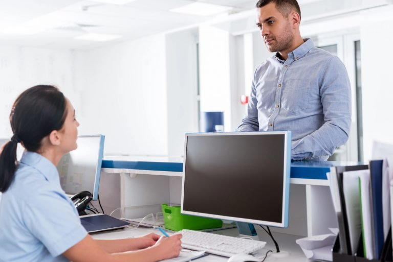 Homem em pé conversa com uma recepcionista sentada atrás de um balcão. Ela veste camisa azul-clara e escreve em um papel. À frente dela há computador, teclado e materiais de escritório. O ambiente é claro e tem aparência de recepção de empresa ou instituição.