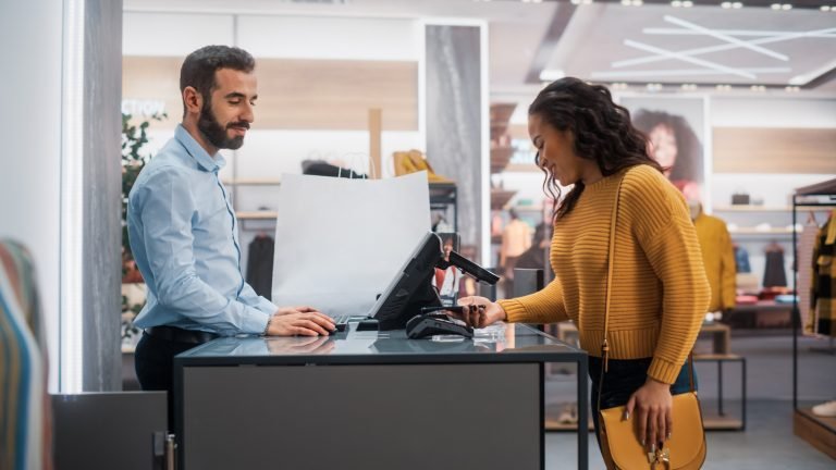 Loja de Roupas: Jovem no Balcão compra roupas de assistente de vendas de varejo amigável, pagando com smartphone NFC sem contato Touch Terminal