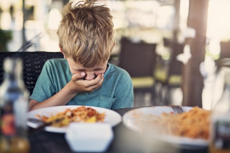 Menino pequeno, seletivo para comer, em um restaurante. Ele não quer comer e chega a sentir ânsia de vômito só de olhar para o prato.